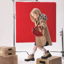 Малко момиче в бежово зимно палто и леопардов шал, държи червена чанта и позира пред червен студиен фон